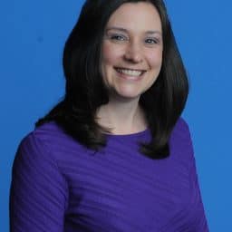 White woman with dark hair, wearing a dark purple sweater against a blue background