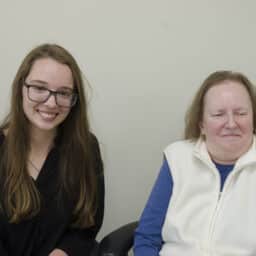 Two women sitting next to each other. The woman on the left is wearing glasses and black shirt. The woman on the right is wearing a white vest and blue shirt.