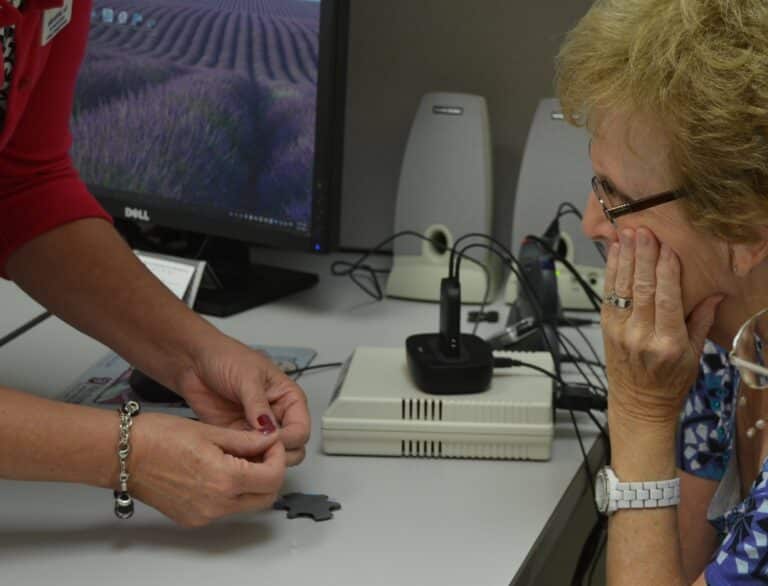 A woman with painted finger nails is showing hearing aid to a Caucasian woman with blonde short hair, wearing glasses.  