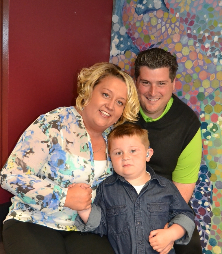 A white woman in a flowered top to the left, a white man wearing a black sweater vest and green top to the right and a white boy wearing a black shirt in the middle.  