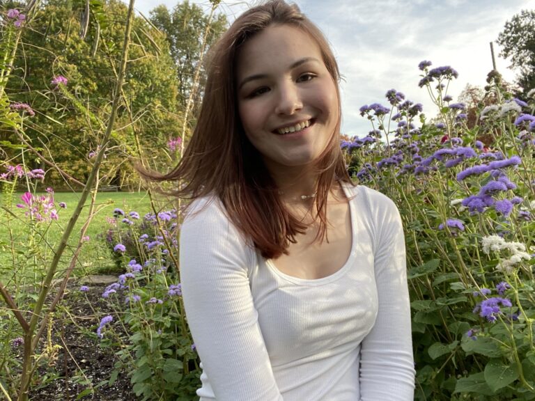 A young woman wearing a white top backed by a field of purple flowers.