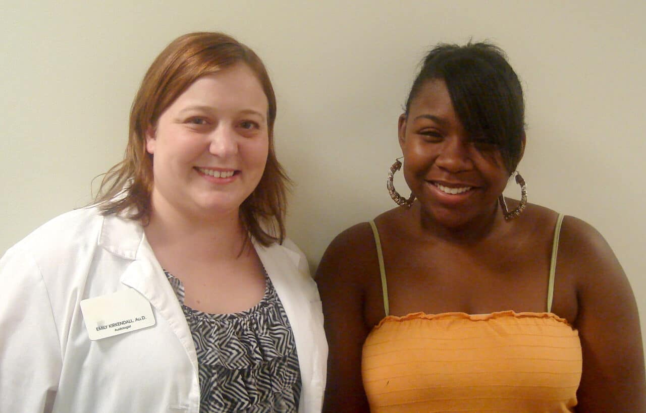 a Caucasian woman in a white lab coat smiling next to a young African American woman wearing an orange tank top.