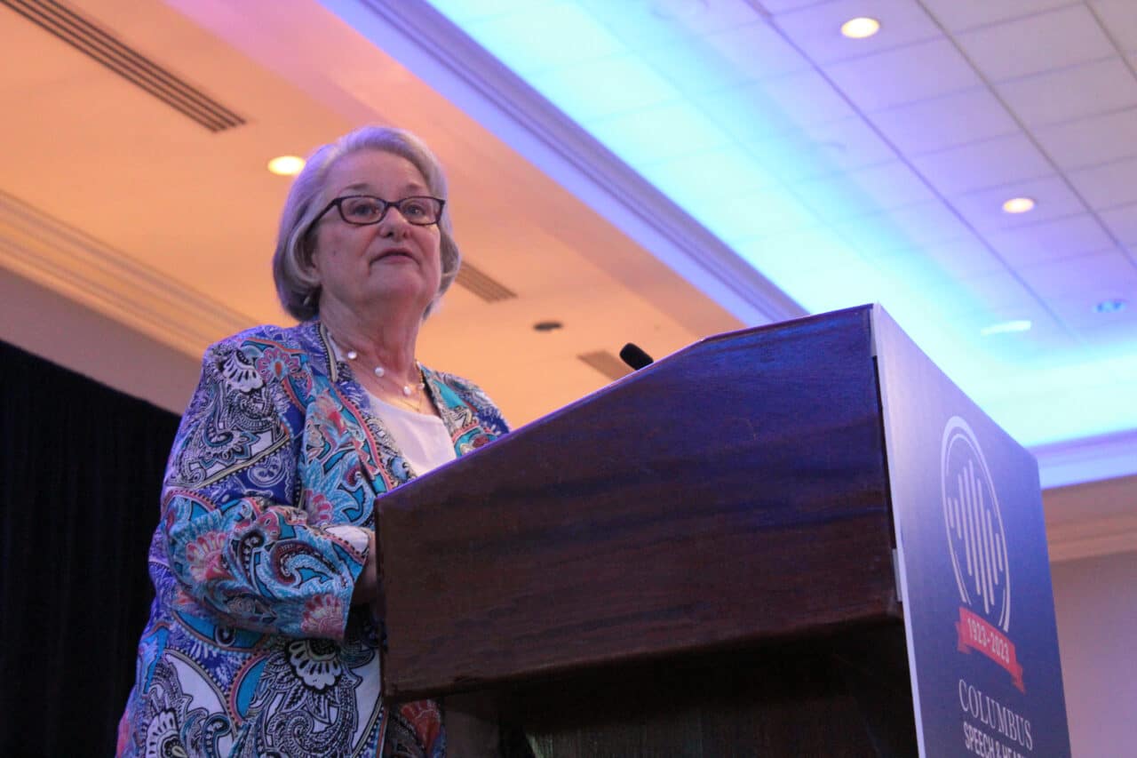 A caucasian woman with short gray hair, in a paisley patterned top, standing a a podium.  